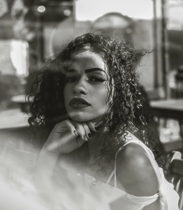 a black and white photo of a woman sitting at a table, pexels contest winner, black curly hair, ariel perez, pout, sitting in a cafe