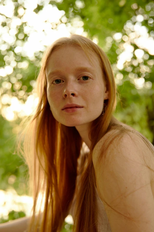 a woman with long red hair sitting on a bench, pale glowing skin, with a tree in the background, julia hetta, close-up portrait film still