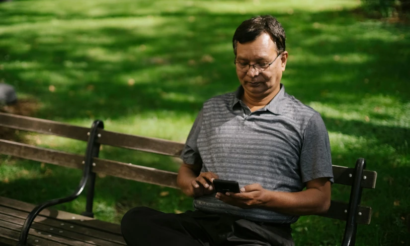 a man sitting on a bench using a cell phone, ranjit ghosh, at a park, digital health, indigenous man