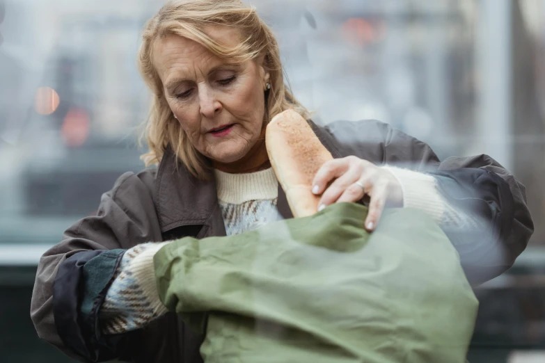 a woman holding a bag full of food, a picture, by Matija Jama, pexels contest winner, she is wearing a wet coat, dementia, holding a baguette, touching her clothes