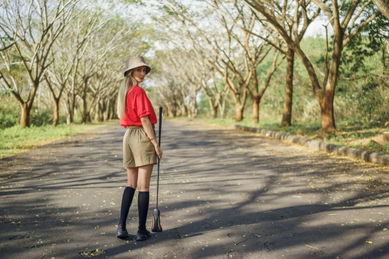 a woman standing in the middle of a road, wearing golf shorts, botanic garden, red shirt brown pants, forest hunter lady