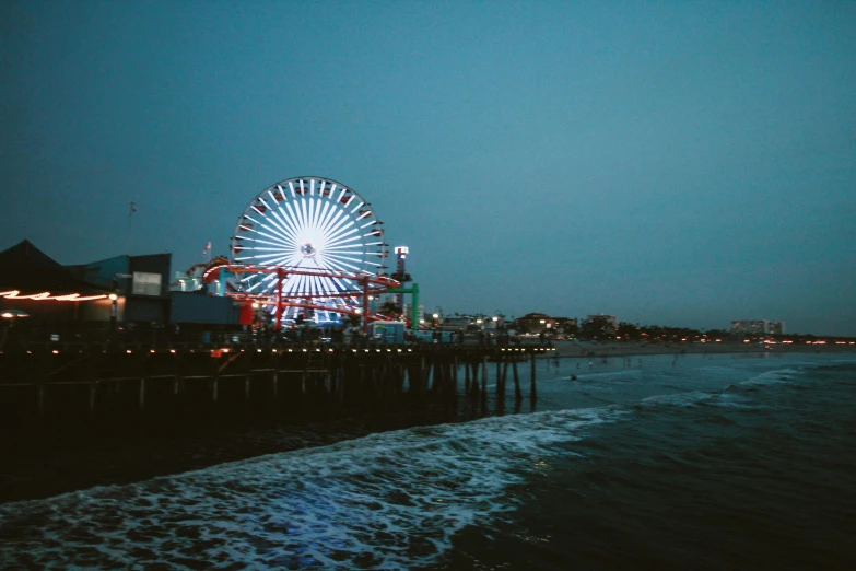 a ferris wheel sitting on top of a pier next to the ocean, inspired by Elsa Bleda, unsplash contest winner, los angeles at night, amusement park buildings, 2000s photo, ☁🌪🌙👩🏾