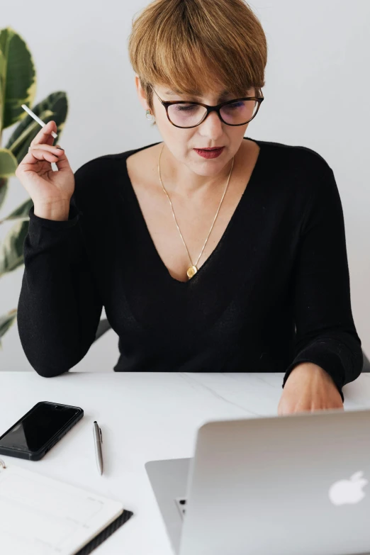 a woman sitting at a table with a laptop and a cigarette, trending on pexels, wearing business casual dress, wearing black glasses, pondering, flatlay