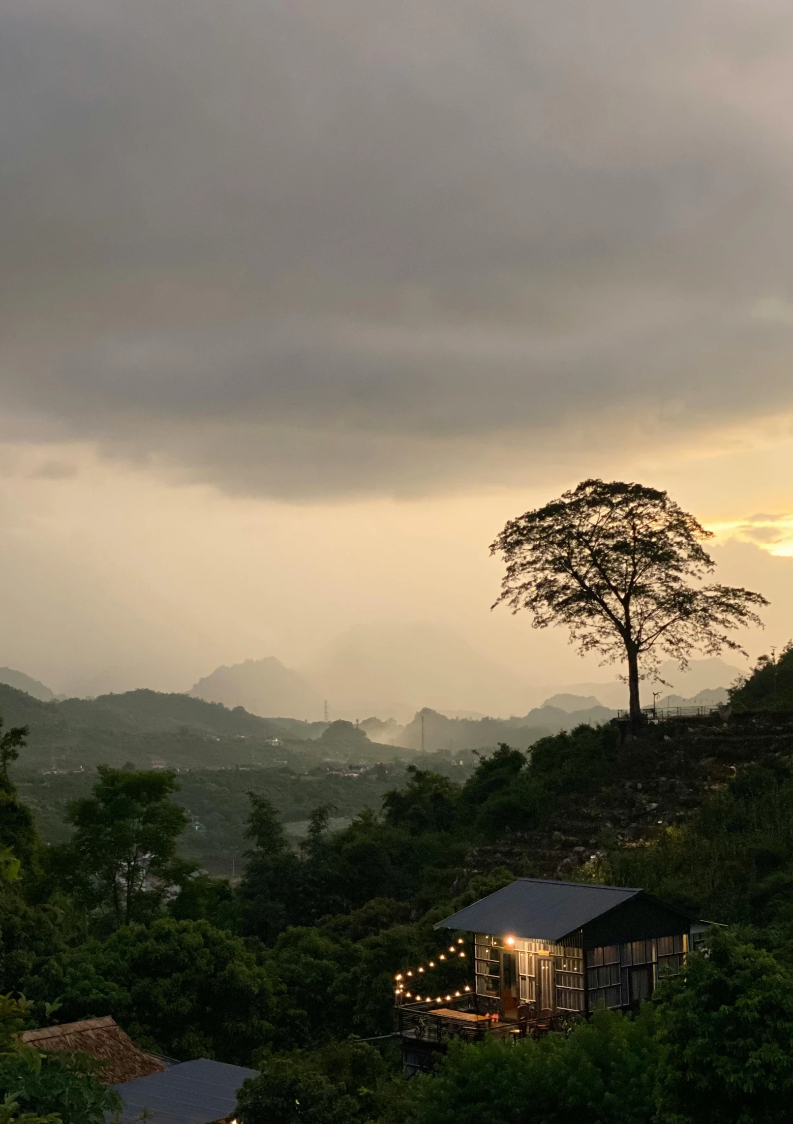 a house sitting on top of a lush green hillside, by Reuben Tam, sumatraism, the bodhi tree at sunset, grey, natural overcast lighting, vietnam