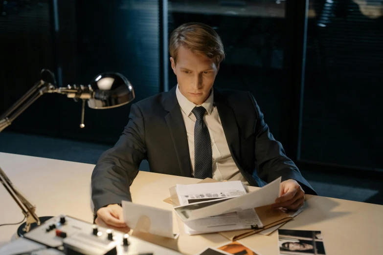 a man sitting at a desk reading a book, inspired by Harry Haenigsen, pexels contest winner, hyperrealism, wearing a suit, film still promotional image, mid night, papers