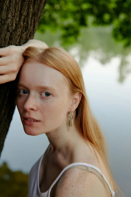a woman leaning against a tree next to a body of water, an album cover, by Grytė Pintukaitė, soft red hair, with a white complexion, close - up portrait shot, naturalist