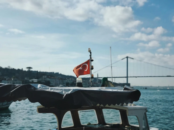 a boat on a body of water with a bridge in the background, by Niyazi Selimoglu, pexels contest winner, hurufiyya, red and black flags waving, 🦩🪐🐞👩🏻🦳, 2. sigara aşırmak, with a white
