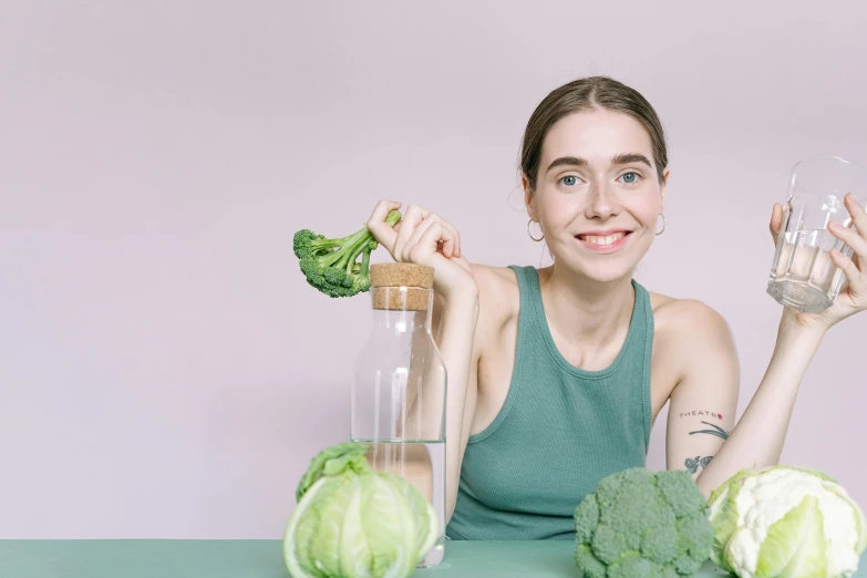 a woman sitting at a table holding a glass of water, with broccoli hair, pale green halter top, background image, confident holding vegetables