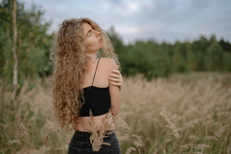 a woman standing in a field of tall grass, inspired by Elsa Bleda, trending on pexels, renaissance, no makeup wavy hair, wearing a cropped black tank top, with a curly perm, long ashy hair | gentle lighting