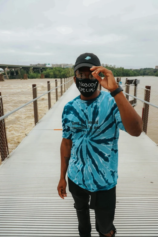 a man wearing a face mask standing on a bridge, an album cover, by Anthony S Waters, trending on unsplash, wearing a tie-dye t-shirt, teal headband, black man, wearing sunglasses and a hat