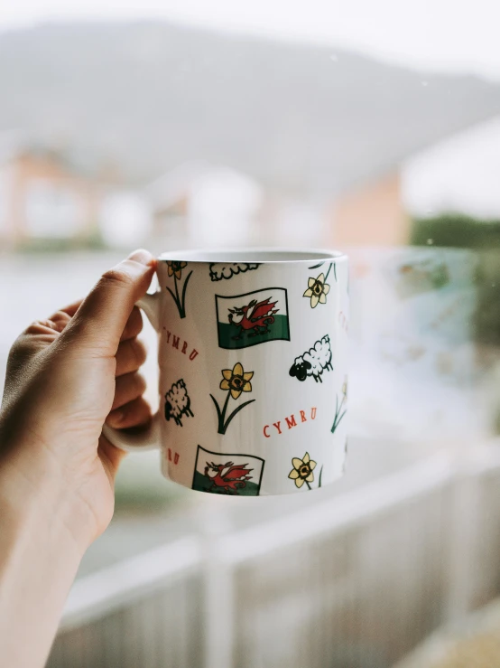 a person holding a coffee cup in front of a window, inspired by Bedwyr Williams, unsplash, arts and crafts movement, welsh flag, patchy flowers, full colour print, side front view