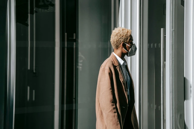 a man walking down the street wearing a face mask, by Emma Andijewska, pexels contest winner, afrofuturism, short blonde afro, wearing a worn out brown suit, leaning on door, people at work