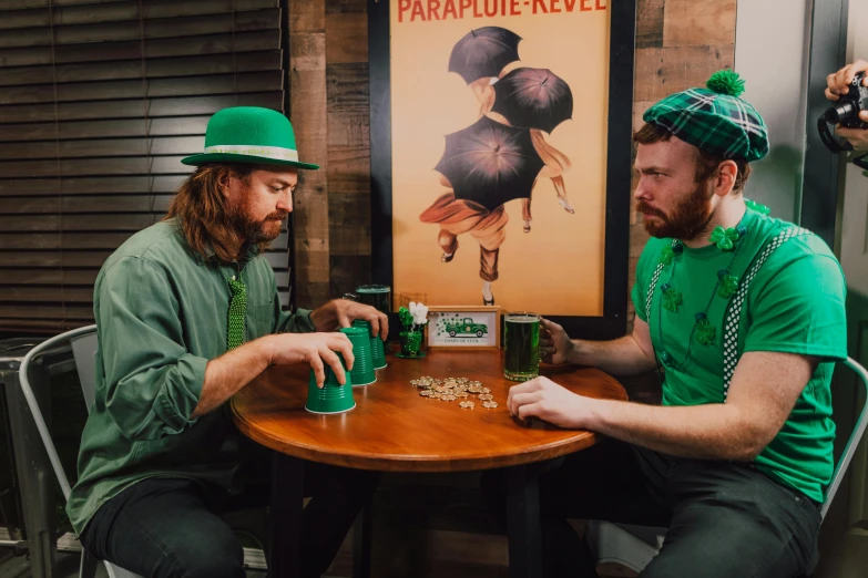 two men sitting at a table playing a game, by Meredith Dillman, pexels contest winner, wearing green tophat, clover, promotional image, pouring