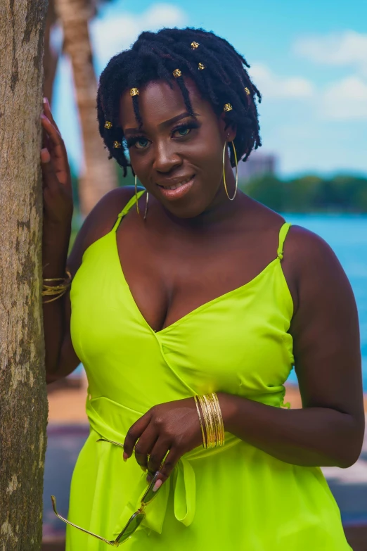 a woman standing next to a tree near a body of water, by Lily Delissa Joseph, wearing a neon green dress, dark skin tone, wearing yellow croptop, close up half body shot