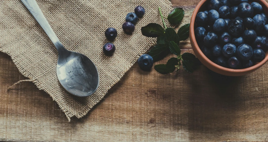 a wooden table topped with a bowl of blueberries, a still life, trending on unsplash, background image, spoon, vintage photo