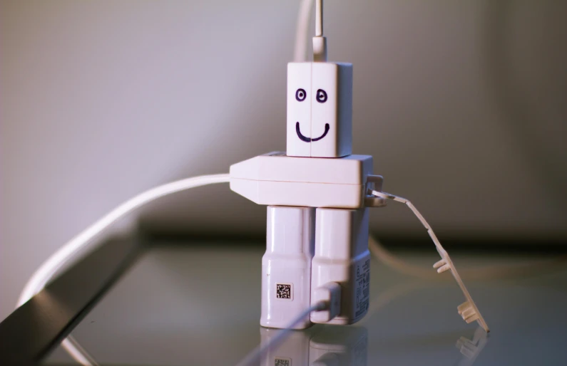 a small white robot sitting on top of a table, charging plug in the chest, smiley face, photograph taken in 2 0 2 0, square