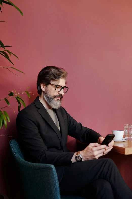 a man sitting at a table using a cell phone, wearing a suit and glasses, small dark grey beard, ignant, mateo dineen