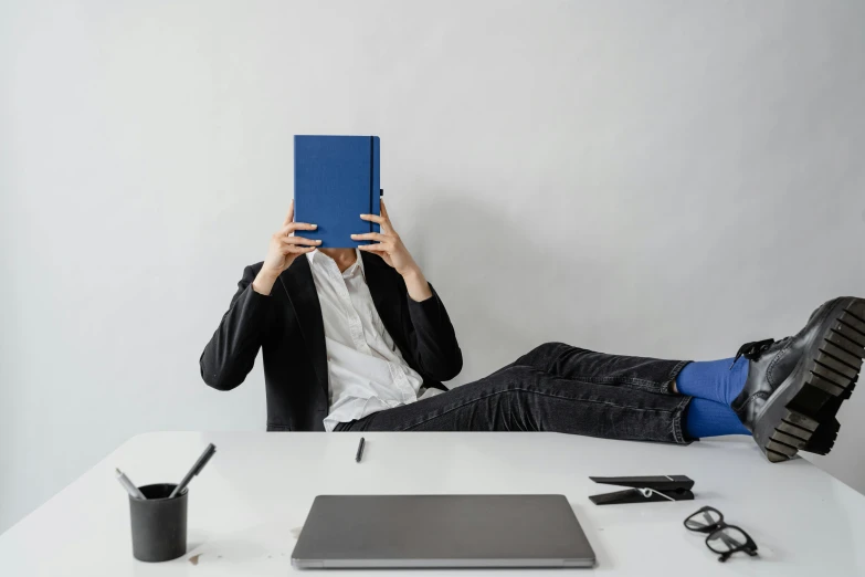 a man sitting at a table covering his face with a book, an album cover, by Paul Bird, trending on unsplash, figuration libre, minimalist home office, blue and black, big googles over her head, office clothes