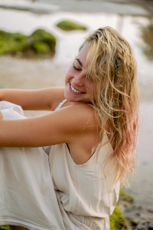 a woman sitting on a beach next to a body of water, happening, turning her head and smiling, blonde flowing hair, smiling laughing, with textured hair and skin