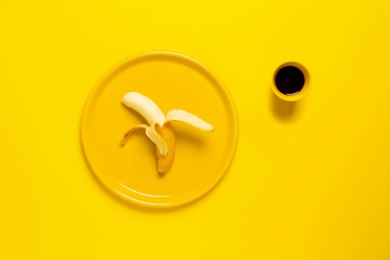 a banana on a plate next to a cup of coffee, by Sven Erixson, pexels contest winner, postminimalism, yellow aureole, made of food, thiago lehmann, 🐿🍸🍋