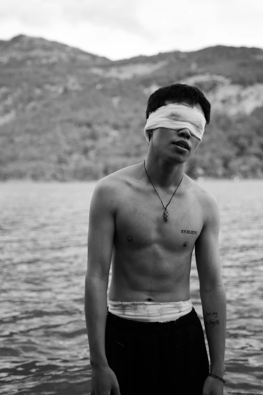 a man with a blindfold standing in the water, a black and white photo, by Caro Niederer, realism, jimin\'s right eyelid swollen, declan mckenna, new song, cracked body full of scars