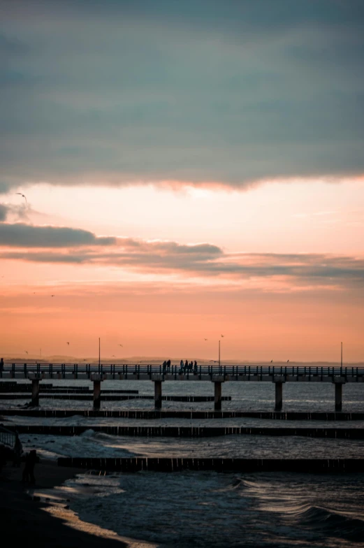 a group of people standing on top of a beach next to the ocean, by Tobias Stimmer, pexels contest winner, bridge, cold sunset, harbor, plain