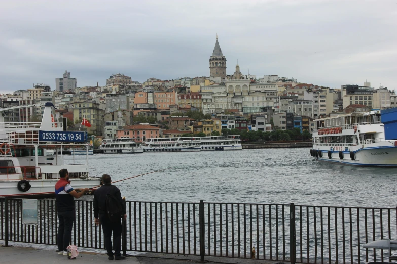 a couple of people standing next to a body of water, a picture, istanbul, buildings in the distance, slide show