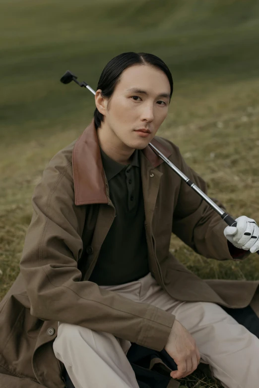 a man sitting on the ground holding a golf club, inspired by Fei Danxu, wearing a brown leather coat, model is wearing techtical vest, darren quach, on a green hill