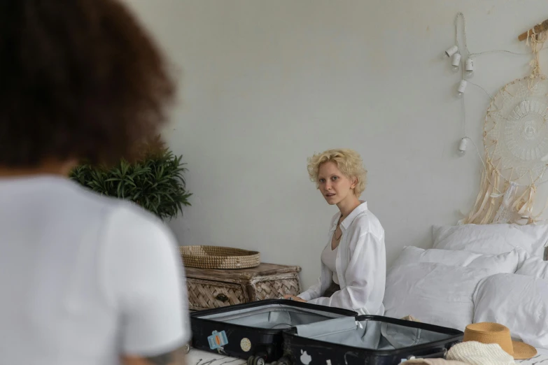 a woman sitting on a bed with a suitcase, by Elsa Bleda, process art, looking in mirror at older self, very very curly blond hair, dressed in a white t shirt, ayahuasca ceremony