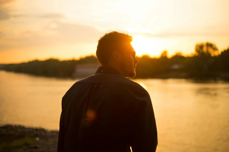 a man standing in front of a body of water, backlit golden hour, thoughtful ), half image, medium-shot