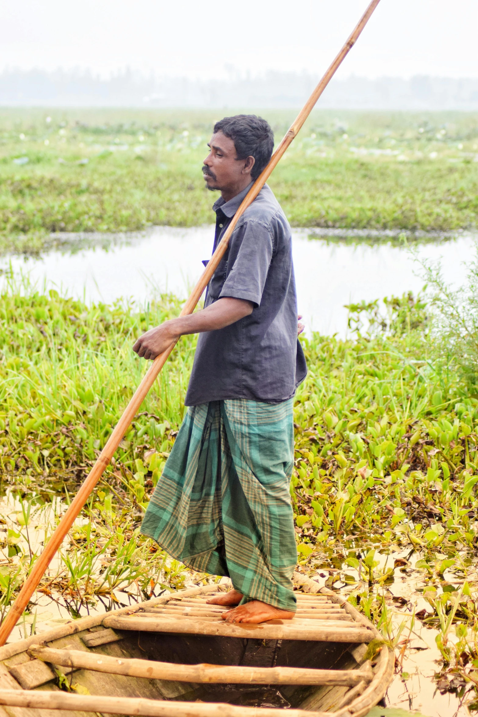 a man standing on top of a wooden boat, by Sunil Das, hurufiyya, walking in high grass field, using a spade, bioremediation, holding a staff