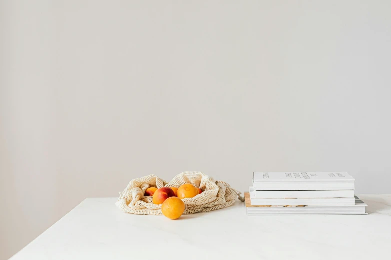 a pile of books sitting on top of a white table, a still life, by Mardi Barrie, unsplash, minimalism, holding mesh bag with bagels, fresh fruit, white marble interior photograph, bowl of fruit