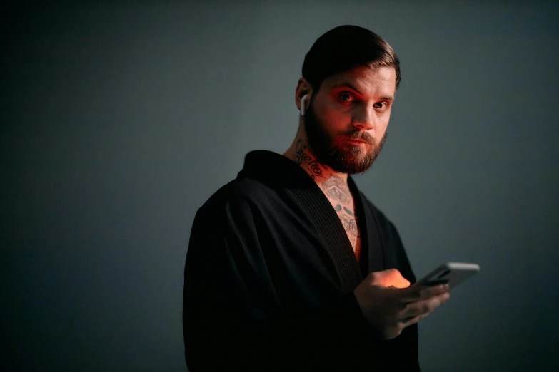 a man in a robe holding a cell phone, a tattoo, by Adam Marczyński, trending on pexels, purism, with a beard and a black jacket, glowing, serious look, ryan dening