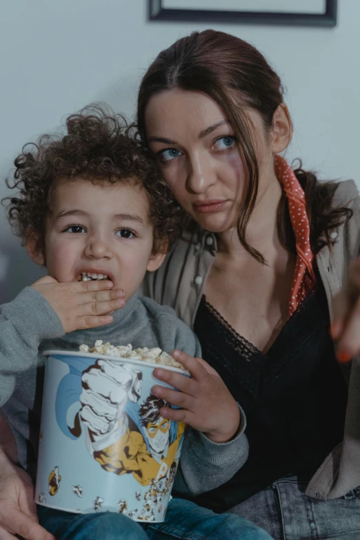 a woman and child sitting on a couch eating popcorn, inspired by Louis Le Nain, trending on reddit, worksafe. cinematic, looking towards the camera, film still promotional image, hunger