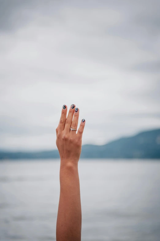 a person holding their hand up in front of a body of water, neat nails, f/8.0, ring, 256435456k film