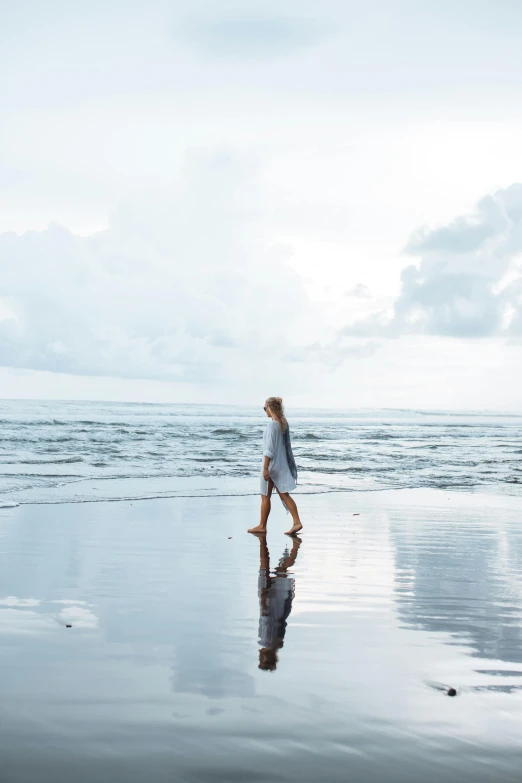 a woman walking on a beach next to the ocean, reflective water, bali, dwell, grey