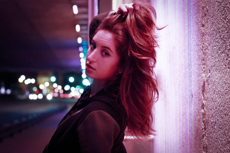 a woman with red hair leaning against a wall, an album cover, inspired by Elsa Bleda, pexels contest winner, photorealism, hero pose colorful city lighting, anna kendrick, purple light, windy hair