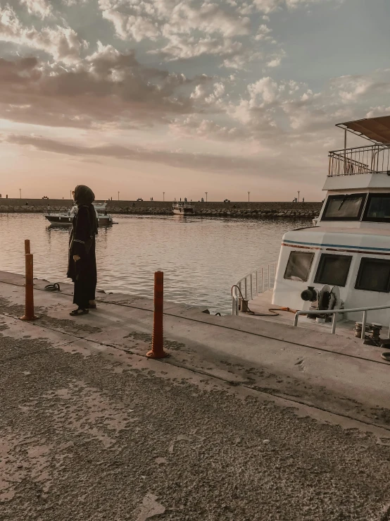 a man standing next to a boat on a body of water, by Ahmed Yacoubi, panoramic view of girl, street photo, cinematic image, facing away