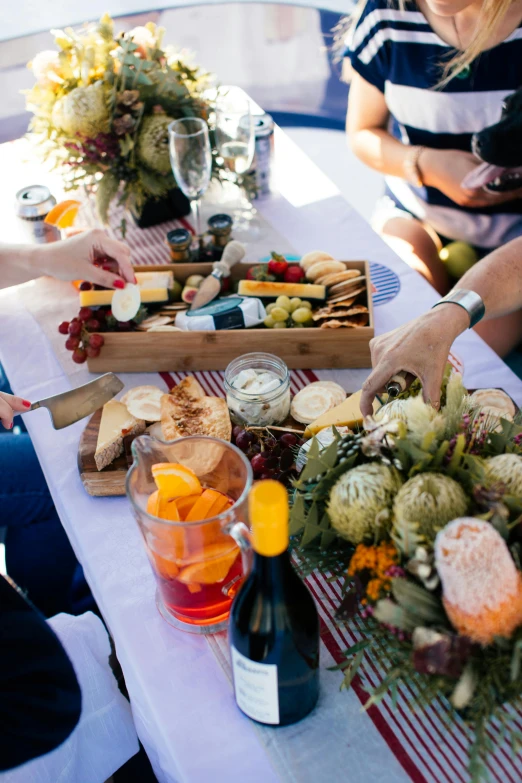 a group of people sitting around a table with food, champagne on the table, eating a cheese platter, sunny day time, harvest