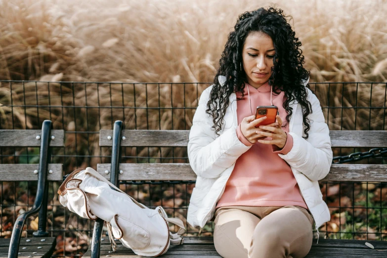 a woman sitting on a bench looking at her cell phone, trending on pexels, happening, curly haired, wearing a pastel pink hoodie, avatar image, indian girl with brown skin