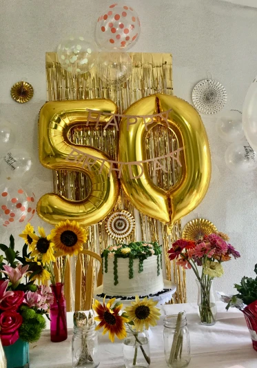 a table topped with a cake covered in gold balloons, 50, bright sun, profile image, flowers