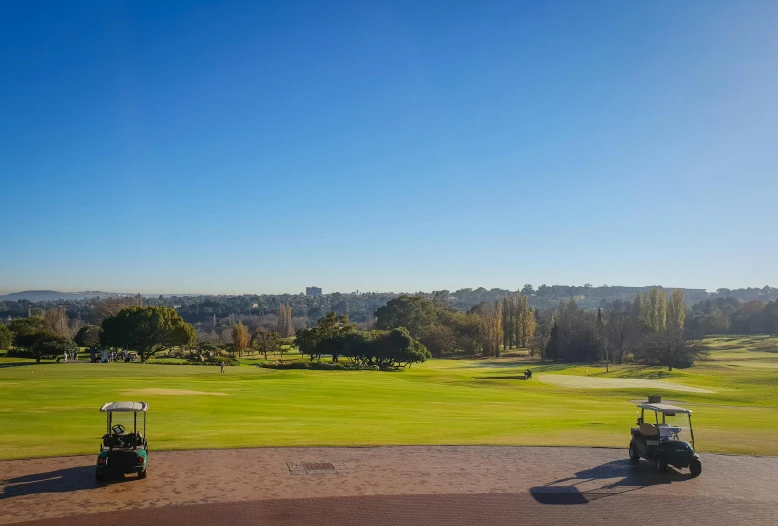 a couple of golf carts sitting on top of a lush green field, with a city in the background, winter sun, green square, crisp image