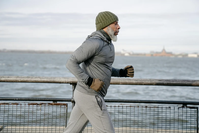 a man running on a boardwalk next to a body of water, he also wears a grey beanie, old gigachad with grey beard, bulging muscles, ap