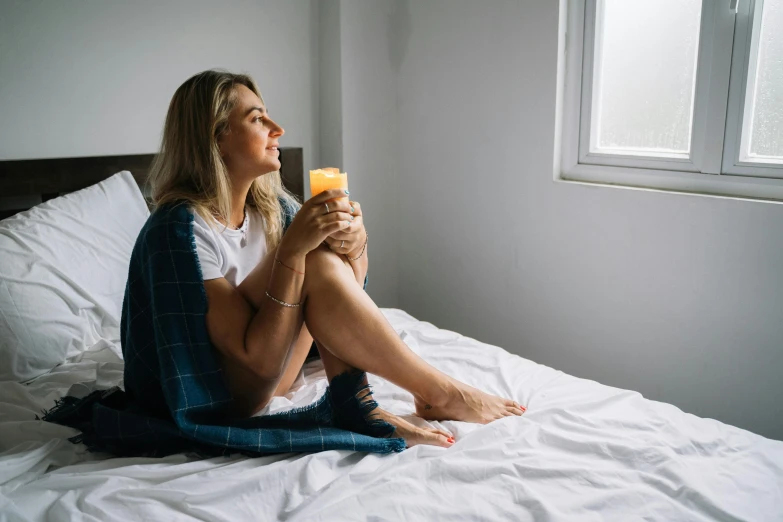 a woman sitting on a bed holding a glass of orange juice, by Carey Morris, pexels contest winner, alana fletcher, looking off to the side, early morning mood, unedited