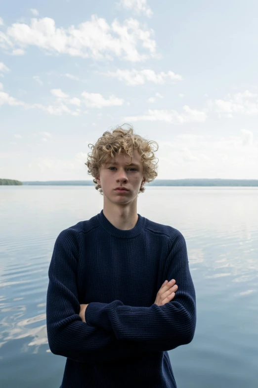 a young man standing in front of a body of water, an album cover, by Attila Meszlenyi, unsplash, renaissance, pale skin curly blond hair, lake house, proud serious expression, finland