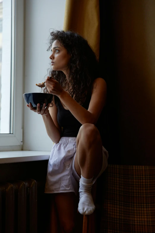 a woman sitting on a window sill eating from a bowl, trending on pexels, renaissance, black, cereal, thoughtful expression, soft glow