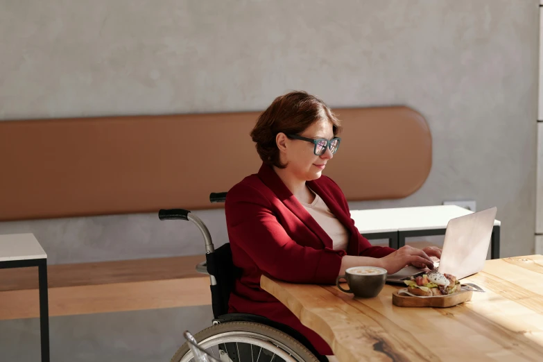 a woman in a wheelchair working on a laptop, a portrait, trending on pexels, hurufiyya, sitting on a mocha-colored table, profile image, eating, wearing a suit and glasses