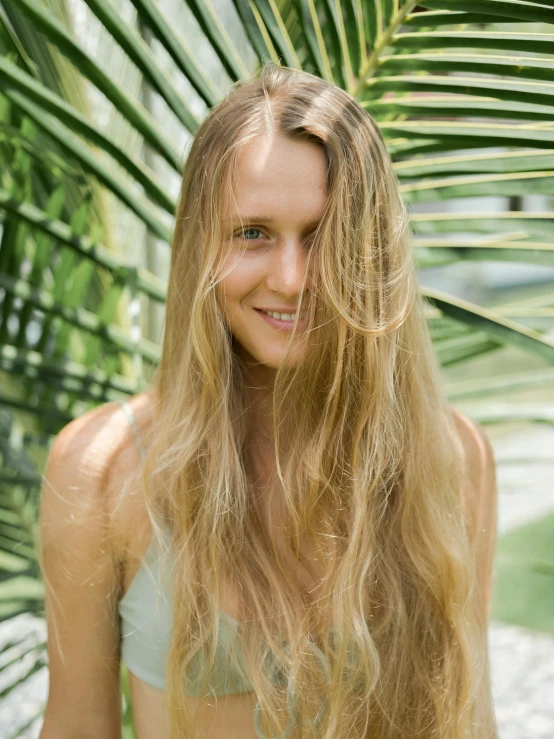 a woman standing in front of a palm tree, by Sara Saftleven, trending on unsplash, light brown long wavy hair, lush greens, translucent skin, smiling slightly