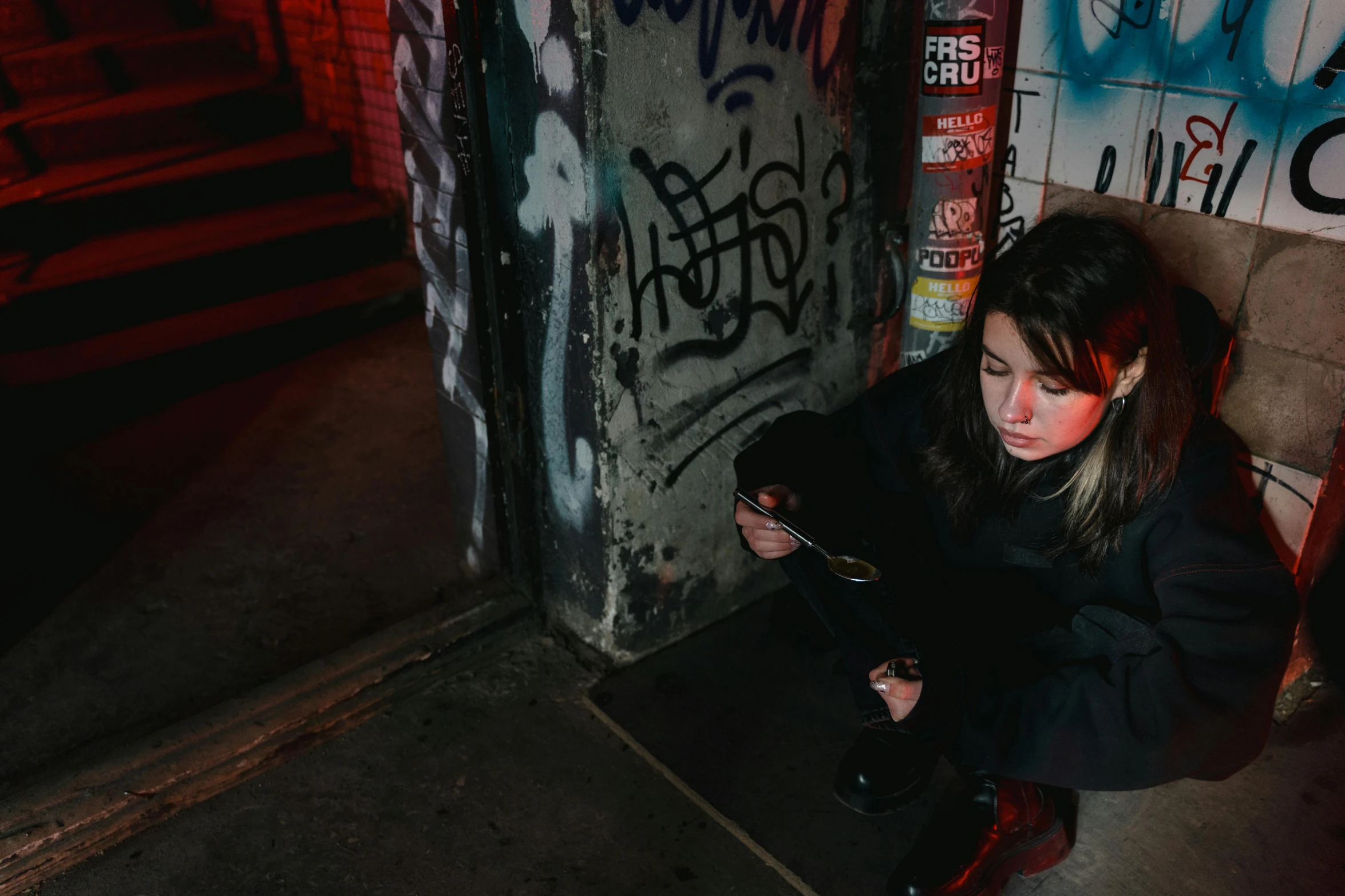 a woman sitting on the ground in front of a wall with graffiti on it, inspired by Elsa Bleda, pexels contest winner, dimly lit underground dungeon, maisie williams, checking her cell phone, young girl