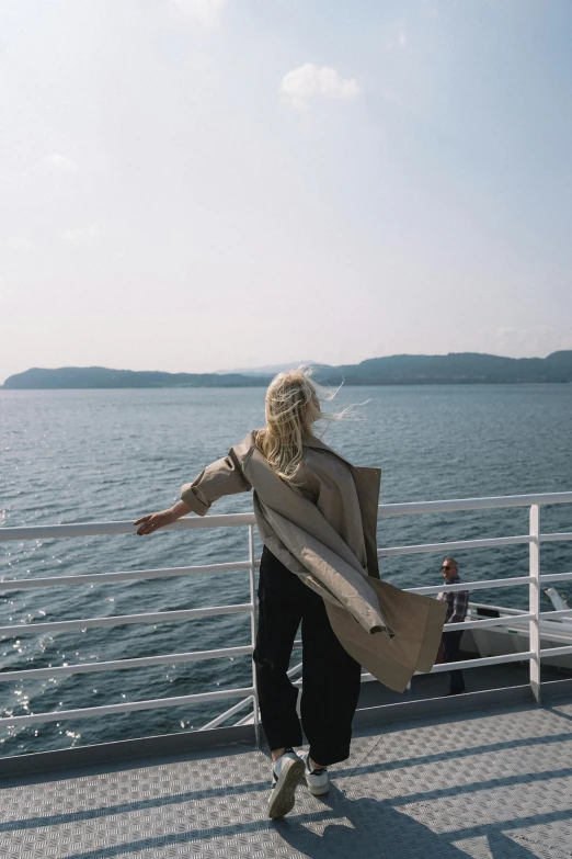 a woman is standing on the deck of a boat, by Niko Henrichon, trending on unsplash, back of emma stone in beige coat, japan shonan enoshima, blonde, horizon view